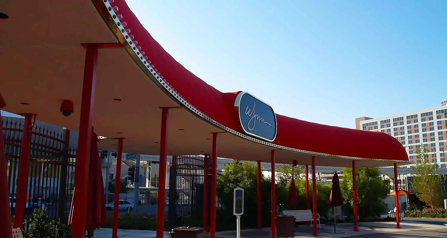 | Las Vegas Awning and StructuresA large red entry canopy provides shade at a hotel entrance, supported by red poles, with a logo on the canopy and greenery around. This is an example of professional awning installation for sun protection in Las Vegas.