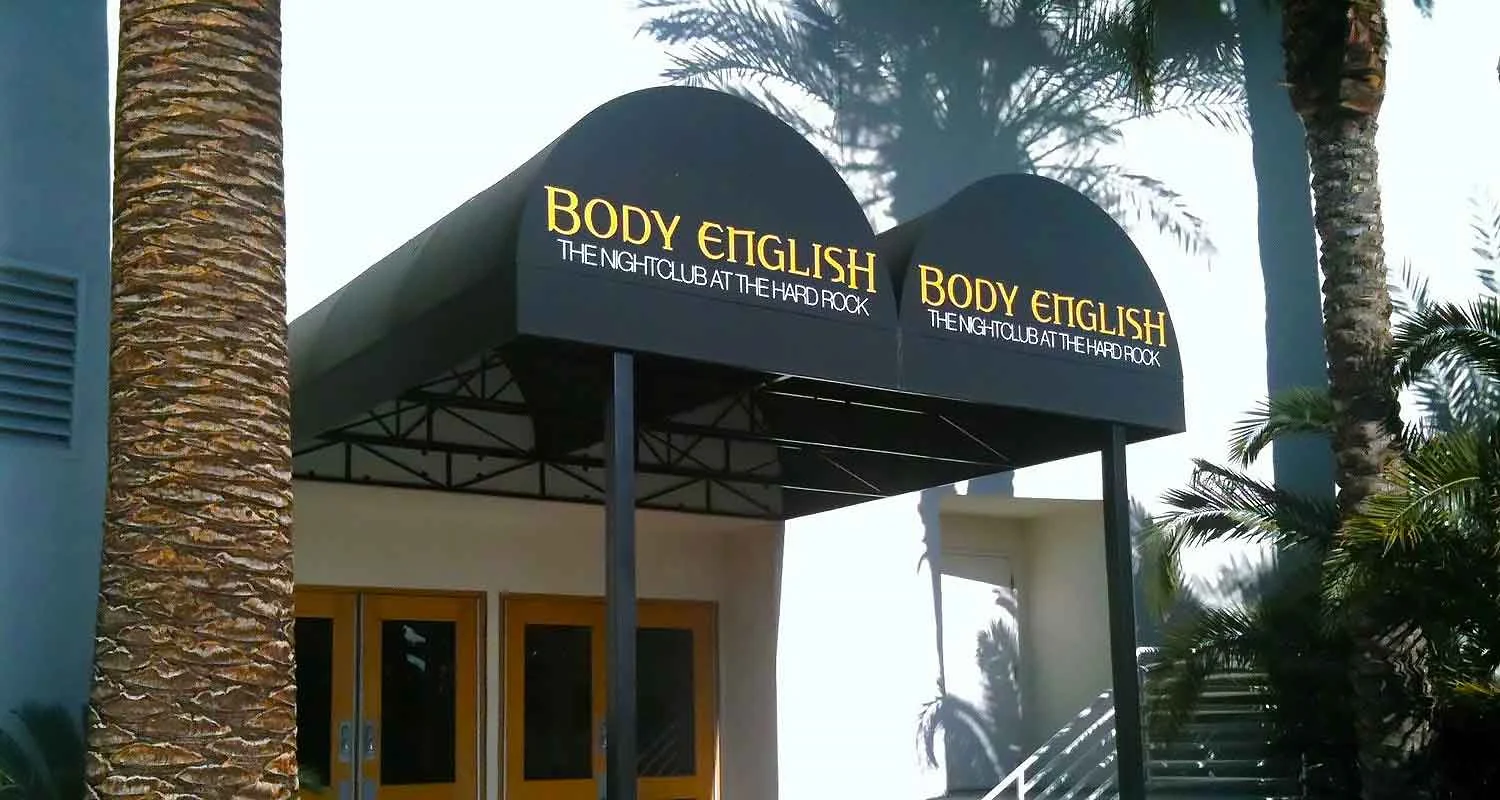 | Las Vegas Awning and StructuresTwo black storefront awnings with yellow text cover the entrance of a nightclub in Las Vegas. Palm trees surround the entrance, providing additional shade and enhancing the aesthetic. These custom awnings offer sun protection and style.